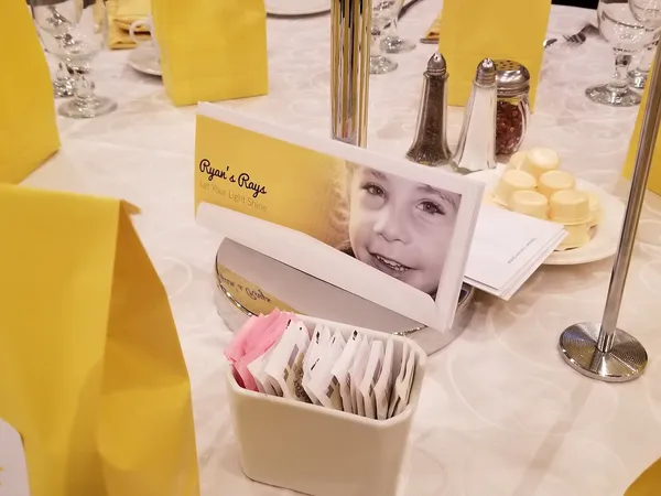 A Ryan’s Rays table card showing a photo of a child, beside a yellow gift bag