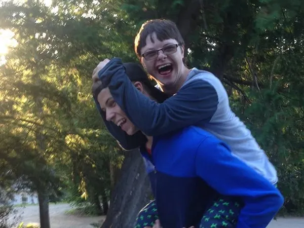 A laughing camper riding piggyback on a counsellor along a wooded path at Muskoka Woods
