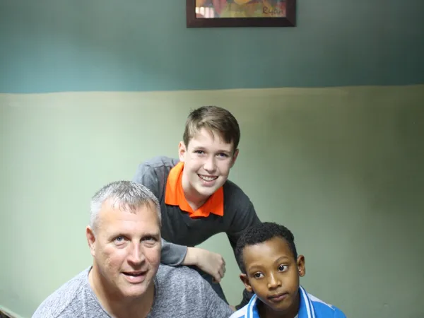 A man and a teenage boy sitting with a young boy in a home, below a framed painting of a smiling child