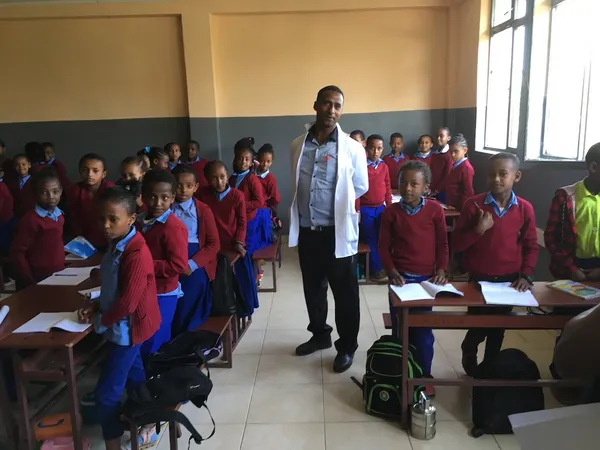 A teacher standing among students in red and blue school uniforms at their classroom desks