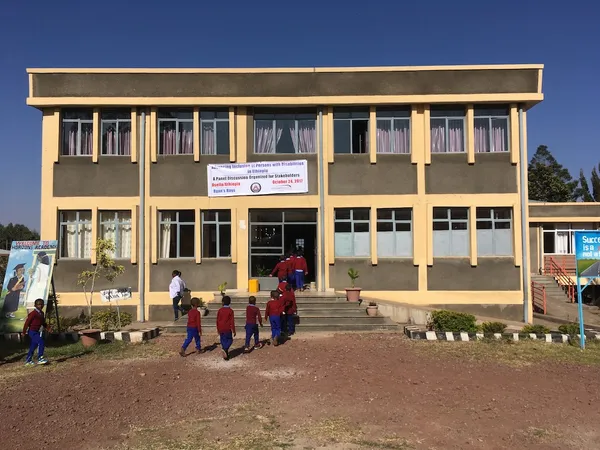 Children walking towards the entrance of a two-storey school building with a banner across the front