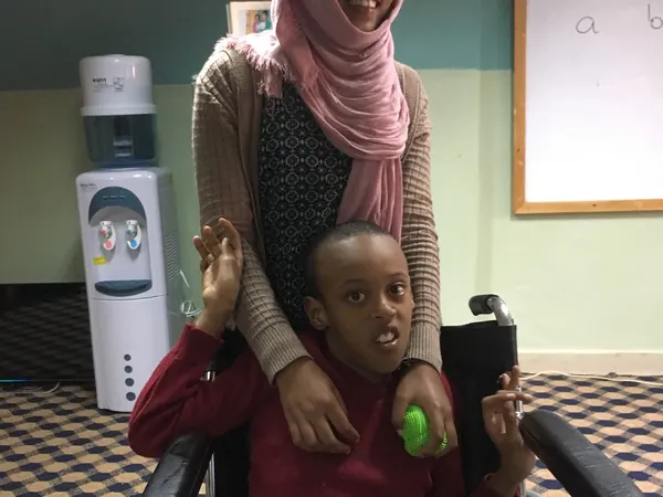A girl in a pink headscarf standing behind a boy seated in a wheelchair in a classroom