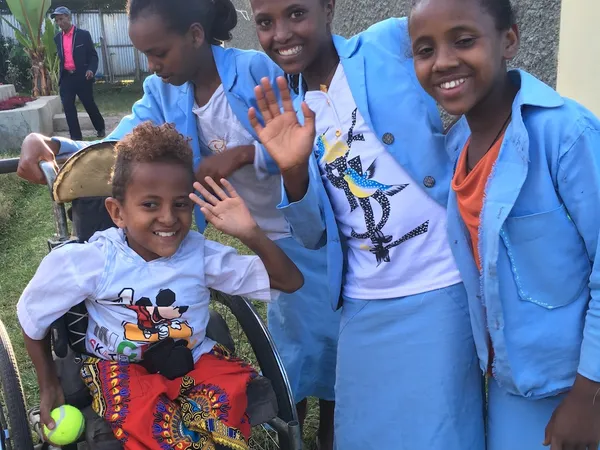 Three girls in blue school uniforms waving beside a younger child seated in a wheelchair