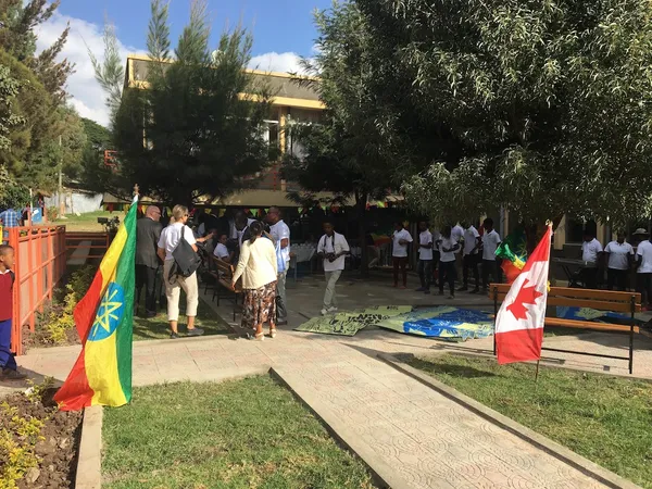 People gathered under trees in a paved school garden, with Ethiopian and Canadian flags flying