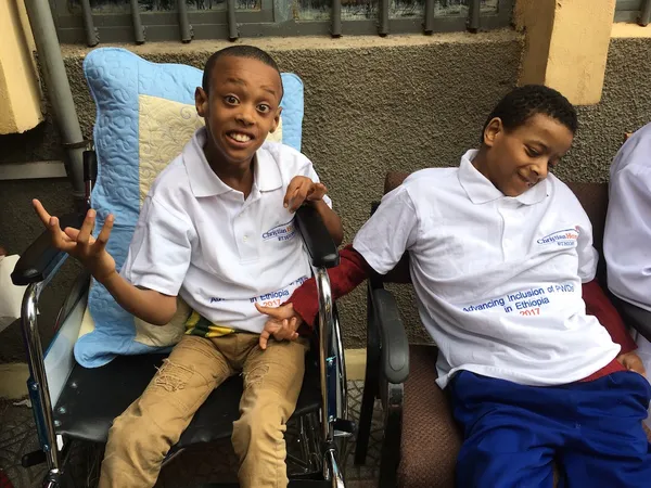 Two boys in white T-shirts, one seated in a wheelchair, smiling on a veranda