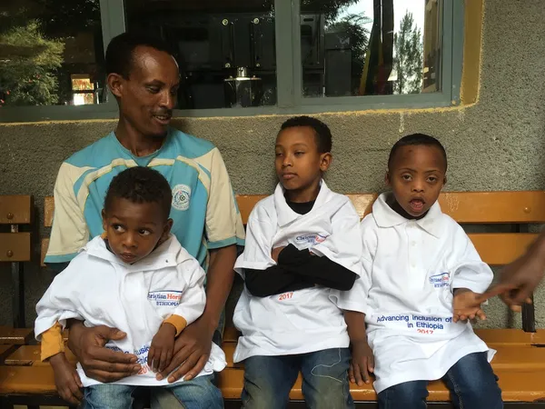A man seated on a bench with three children wearing white project T-shirts